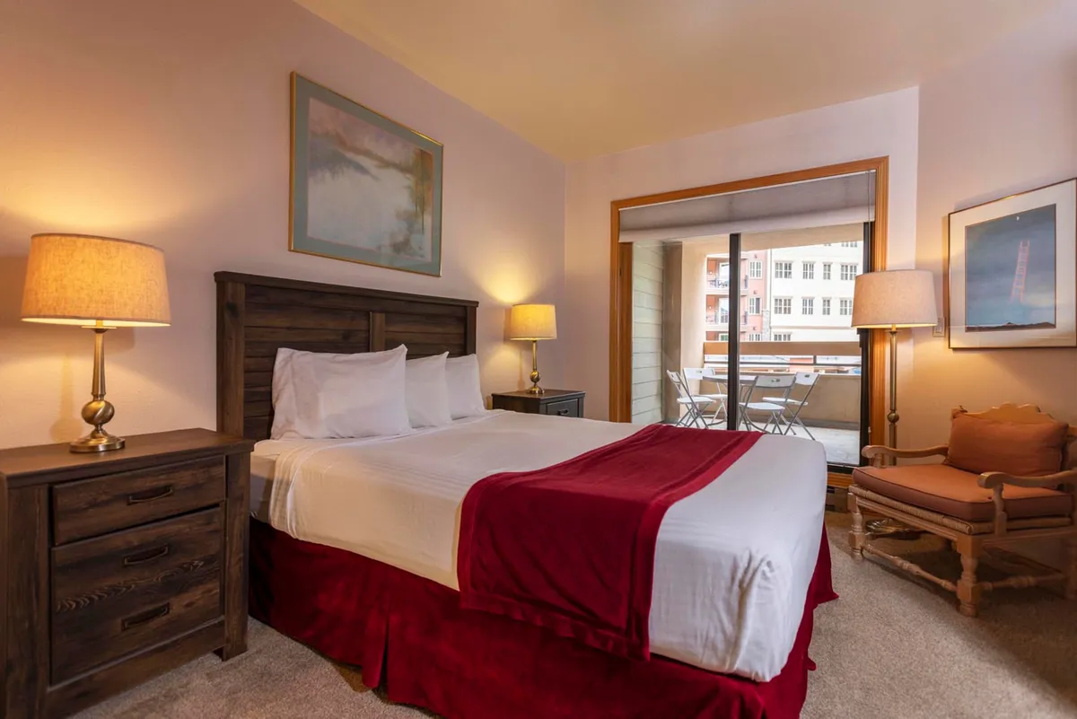 A cozy bedroom with a plush bed adorned with a red throw blanket, nightstands with softly glowing lamps, and direct access to a balcony that reveals mountain views. Natural light streams in, creating a serene and inviting atmosphere ideal for relaxation and restful sleep.