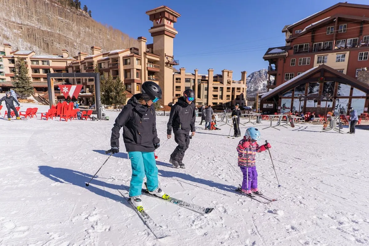 A cheerful family glides down the slopes together beneath bright blue skies, with the resort buildings in the background. The snowy landscape and vibrant winter clothing evoke the excitement of a shared mountain adventure and memorable family moments.