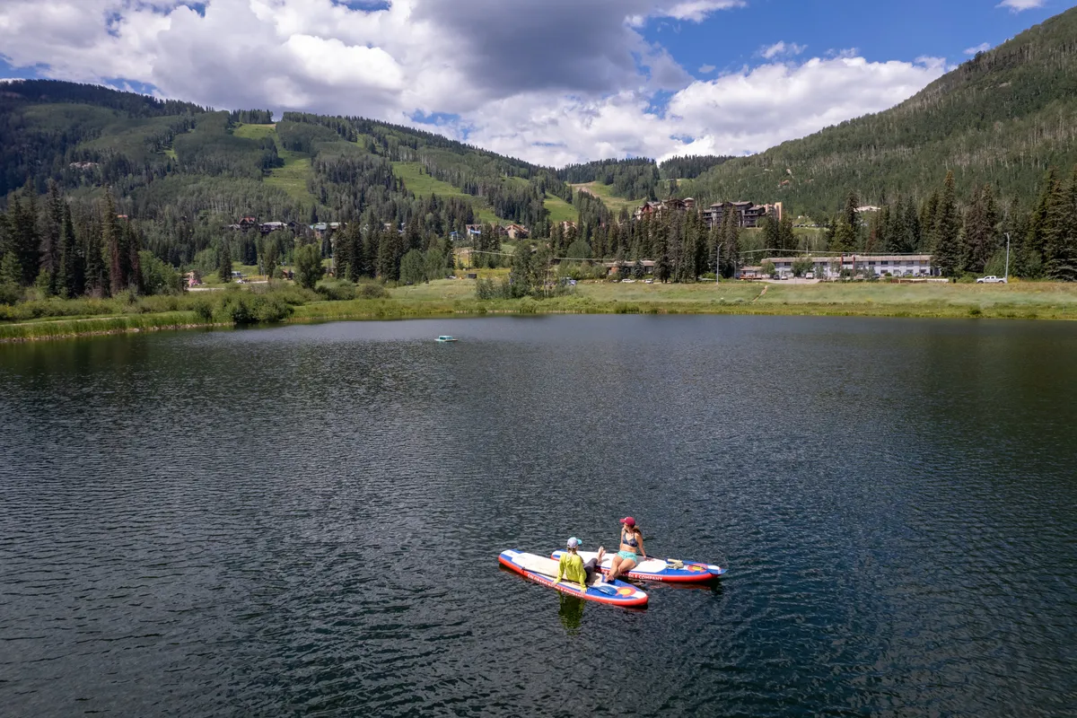 A scenic exterior view featuring two people paddleboarding on a calm mountain lake, surrounded by lush greenery, with majestic mountains in the background. The peaceful setting captures the tranquility and natural beauty of the area, perfect for relaxation and outdoor adventure.