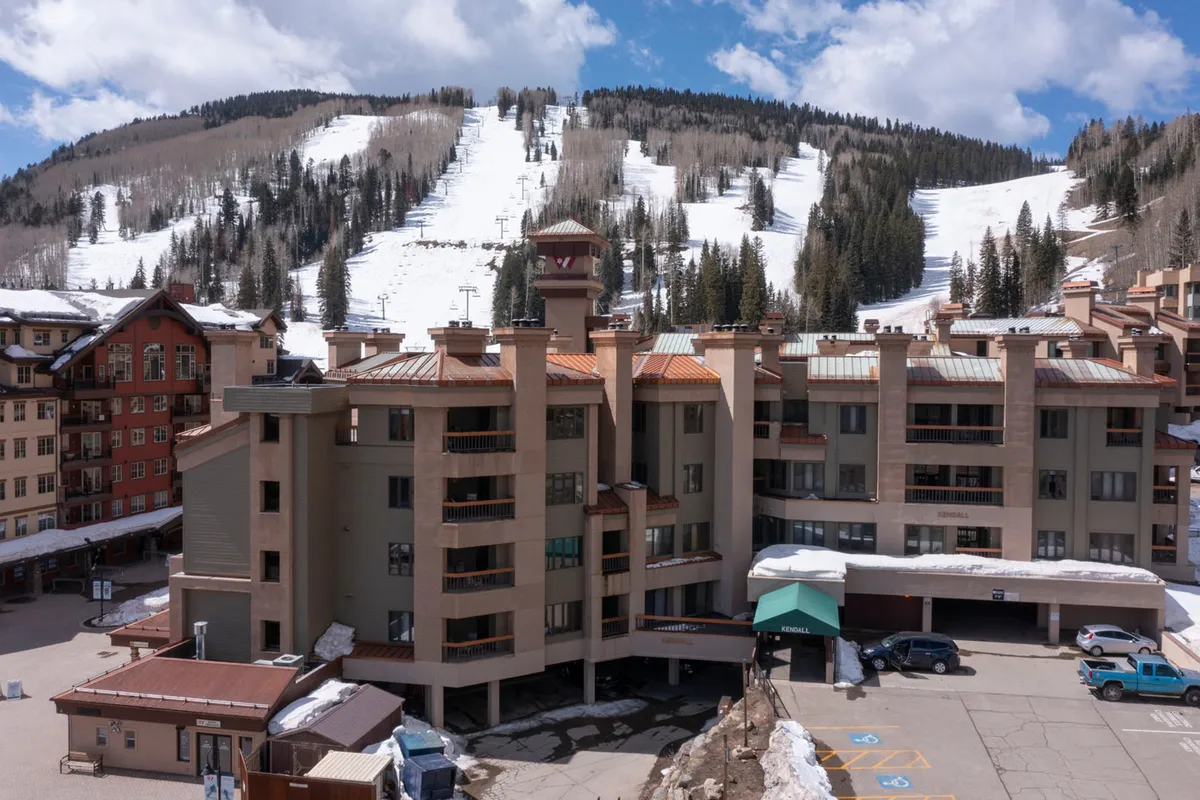 This image captures the exterior of the building with its inviting architecture, the adjacent parking area clearly visible, and the snow-covered slopes forming a scenic mountain backdrop. The overall mood is adventurous and welcoming, suggesting easy transitions between arrival, relaxation, and mountain activities.