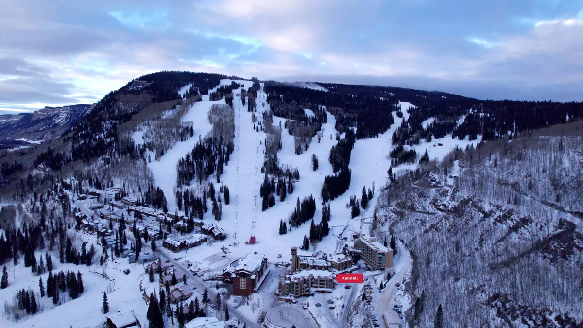 A panoramic view of the ski slopes blanketed in snow, bordered by frosted trees and cozy resort buildings. The scene is set at sunset, casting a serene glow that hints at both active days and relaxing evenings. Hints of ski and snowboard activity convey the vibrant, all-ages fun available just steps from the home base.