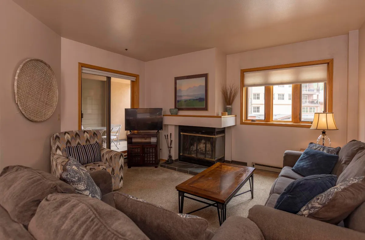 The image showcases a warm and inviting condo living area centered around a glowing wood-burning fireplace, with a plush sofa and cozy coffee table set before expansive windows that invite in mountain light. The arrangement invites guests to unwind, share stories, and savor the comforts of home after a day exploring Durango.