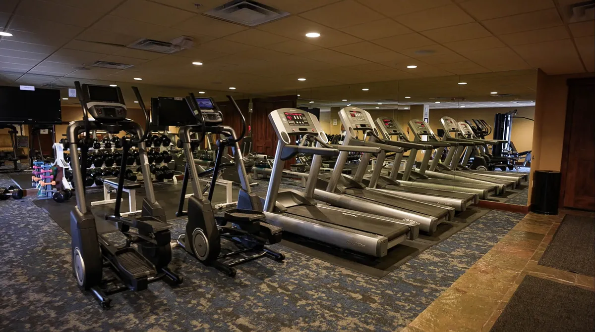 A modern fitness center featuring treadmills, ellipticals, dumbbells, and mirrors, arranged in a bright, motivational environment. The space is clearly designed for both individual workouts and group fitness activities.