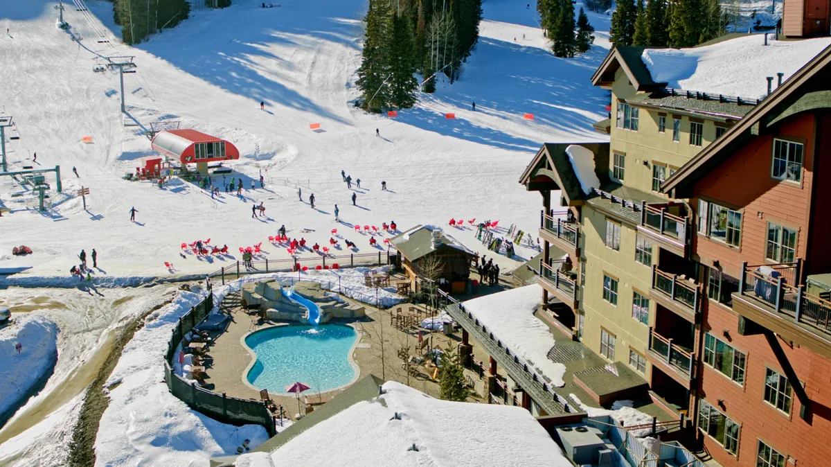 A lively exterior scene showcasing the ski slopes filled with skiers and snowboarders, an inviting outdoor pool with steam rising into the crisp air, and the distinctive lodge architecture of the resort set against a snowy mountain backdrop. The area is bustling with winter activity and relaxation, capturing both adventure and après-ski leisure.