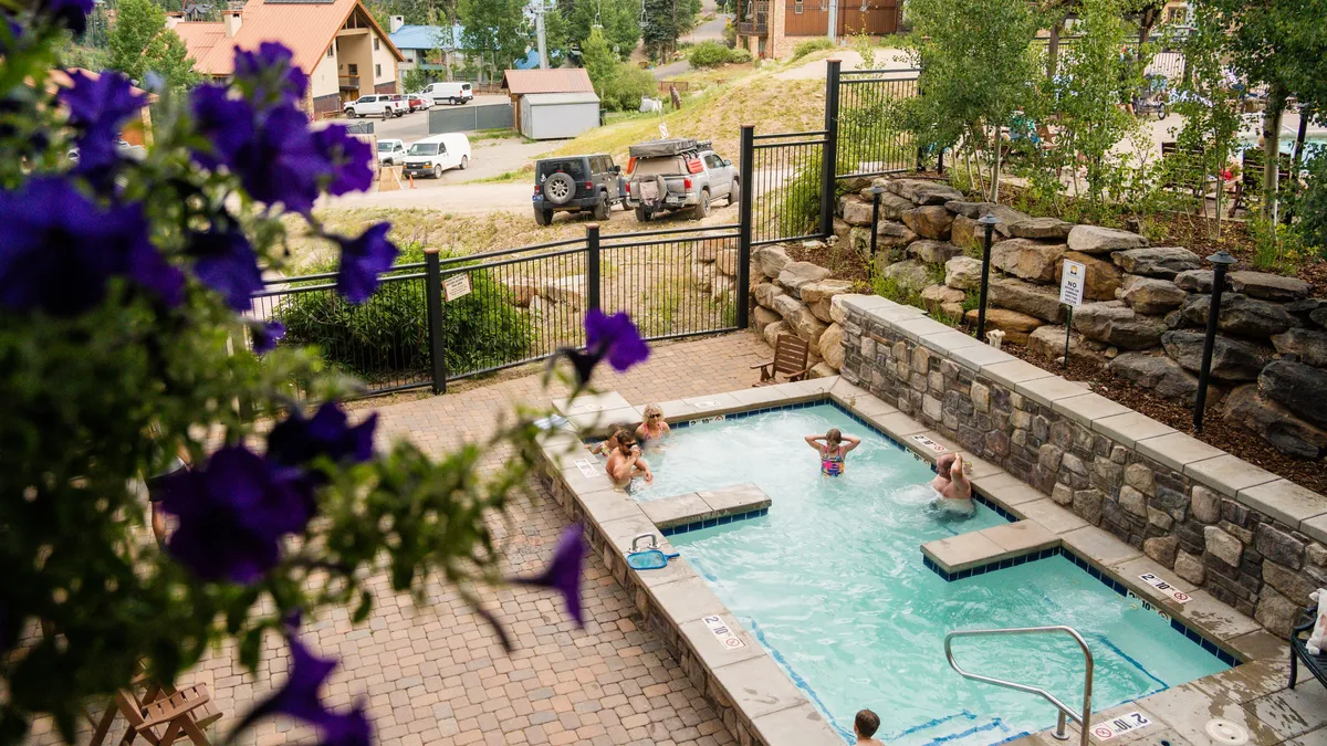 A heated outdoor pool fringed by flowers and greenery, with people swimming, lounging, and socializing. The surrounding landscape adds a resort-like atmosphere, while the pool itself is inviting for play or relaxation.