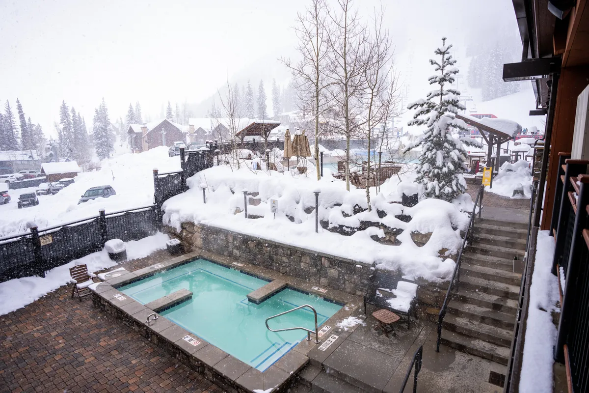 A steaming hot tub set amid a snow-covered landscape, surrounded by trees, with guests relaxing and enjoying the tranquil, wintry scenery. The warm water contrasts with the chilly air, creating a cozy, rejuvenating atmosphere perfect for après-ski relaxation.